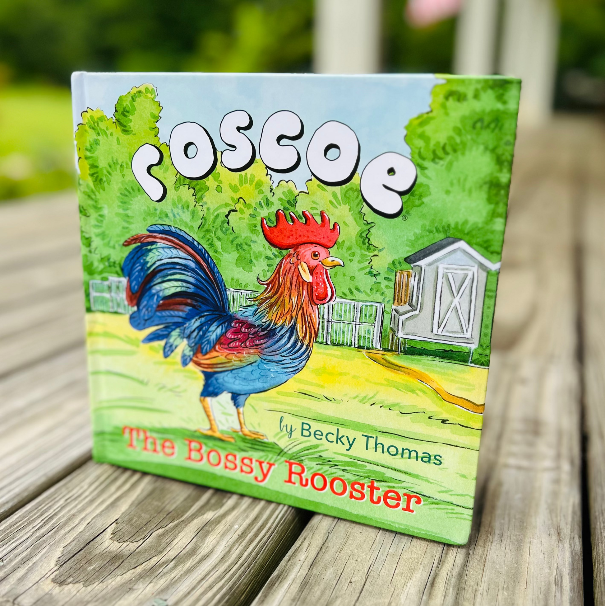 Book titled 'Poscoe the Bossy Rooster' by Becky Thomas on a wooden surface with a blurred background.